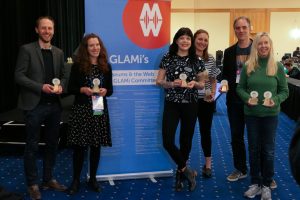 A group of six people stand next to an MW banner and hold their GLAMi Awards. 